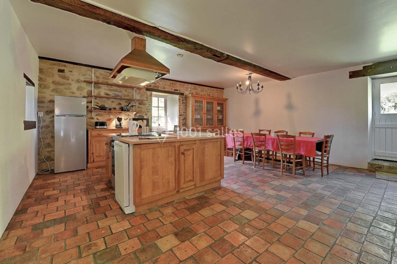 Cuisine spacieuse avec îlot central en bois, table à manger, sol en carreaux de terre cuite et murs en pierre apparente.