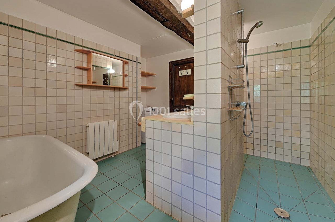 Salle de bain avec baignoire, douche à l'italienne, carrelage beige et bleu, et poutre apparente au plafond.