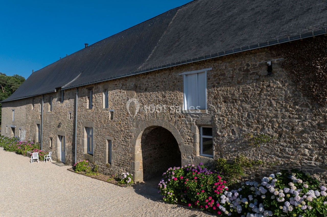 Façade en pierre d'une bâtisse ancienne avec des hortensias colorés et une cour gravillonnée sous un ciel bleu.