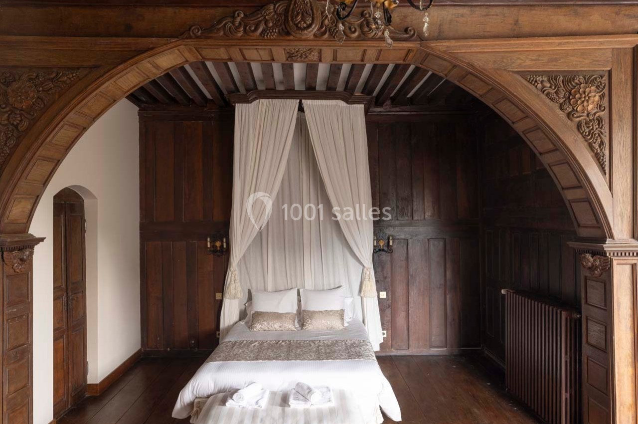 Lit à baldaquin avec draperies blanches dans une chambre aux murs en bois sculpté et parquet ancien.