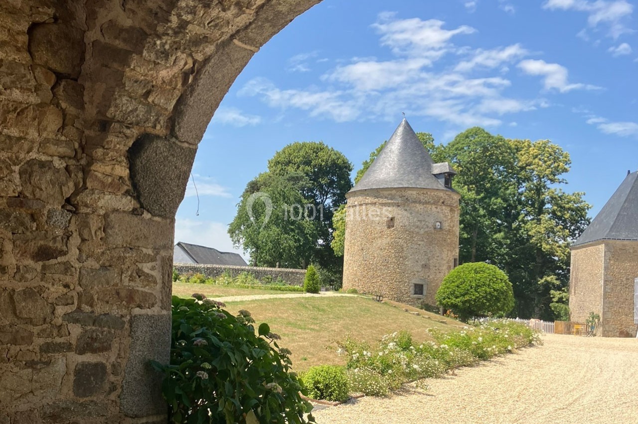 Vue d'une tour en pierre avec un toit conique, entourée de verdure et d'un chemin gravillonné sous un ciel bleu.