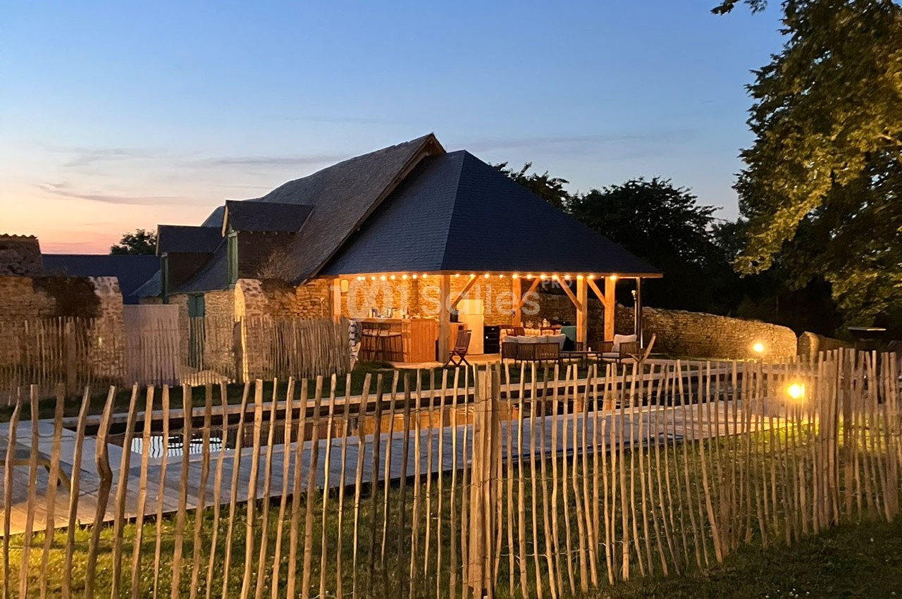 Maison en pierre avec toit en chaume, éclairée le soir, entourée d'une clôture en bois et d'un jardin.