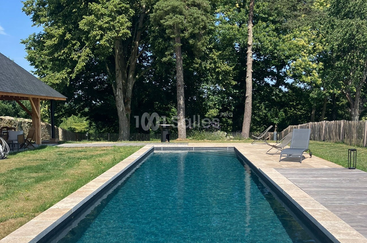 Piscine rectangulaire entourée de pelouse, terrasse en bois et chaises longues, bordée d'arbres en arrière-plan.