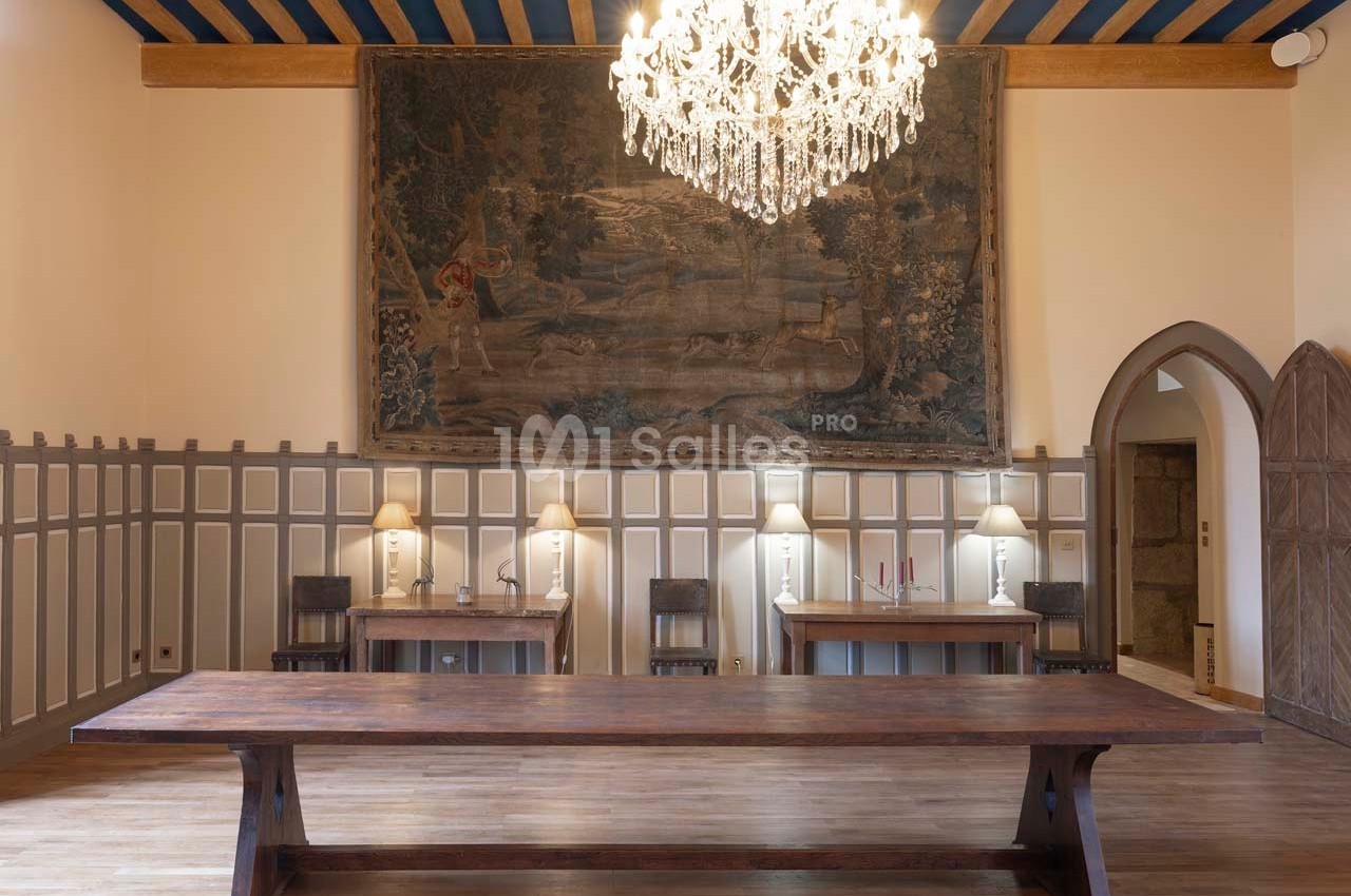 Salle avec une grande table en bois, lustre en cristal, tapisseries murales et éclairage doux par des lampes de bureau.