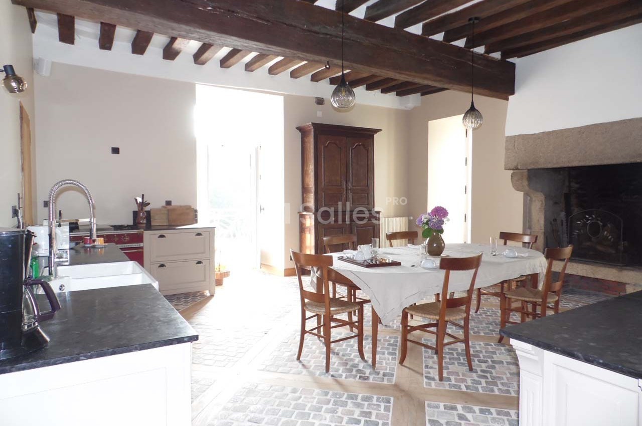Salle à manger lumineuse avec table dressée, poutres apparentes, cheminée en pierre et sol en carreaux de pierre.