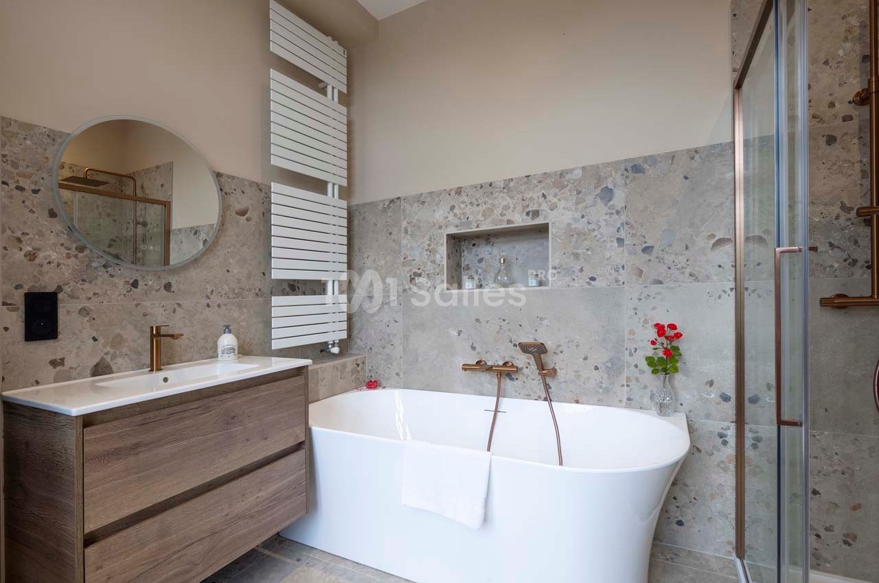Salle de bain moderne avec baignoire blanche, meuble en bois, miroir rond et carrelage effet pierre.