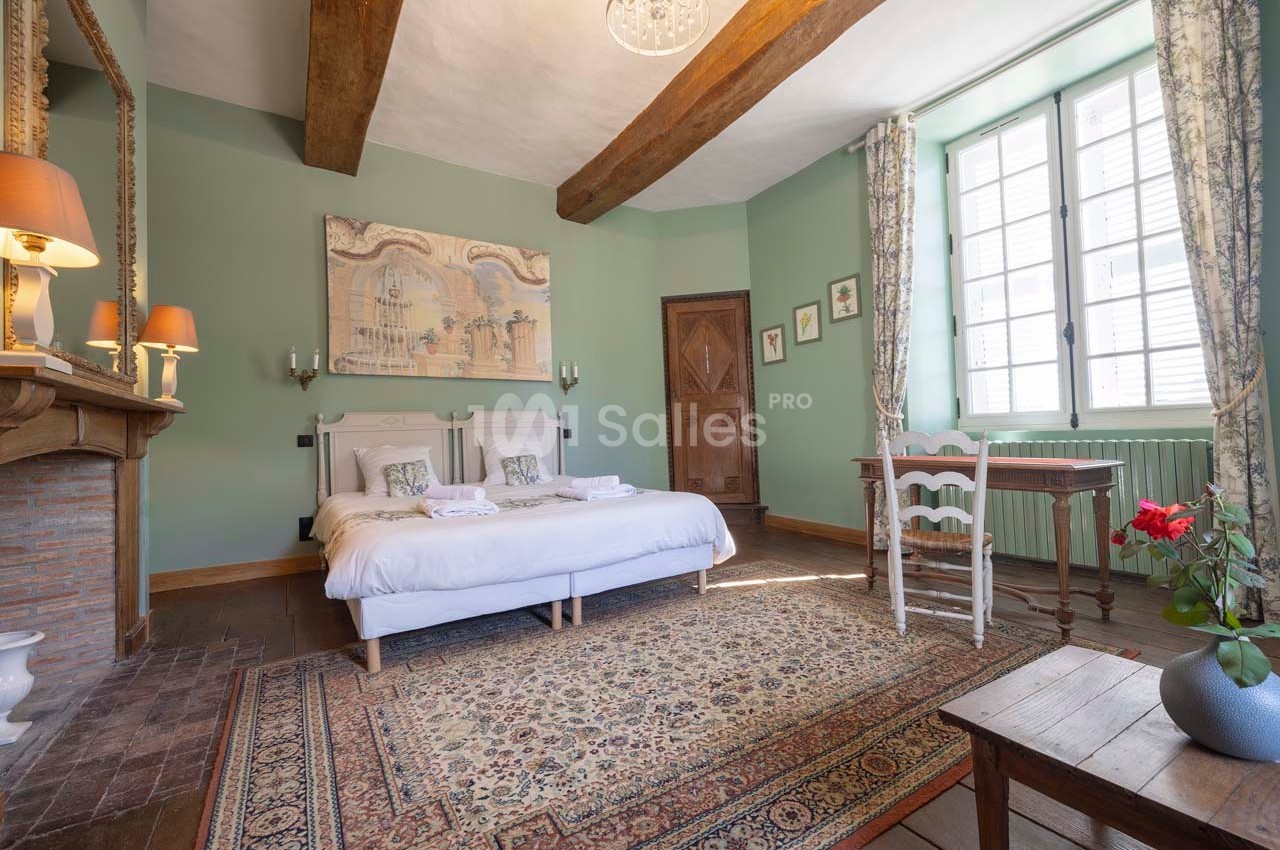 Chambre lumineuse avec lit double, poutres apparentes, murs verts, tapis coloré et mobilier en bois.