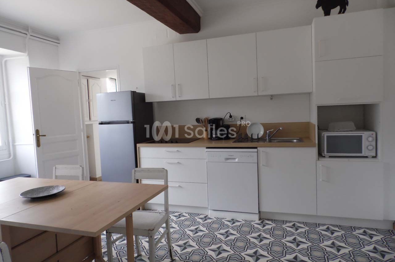 Cuisine lumineuse avec meubles blancs, réfrigérateur, micro-ondes, lave-vaisselle et table en bois sur sol à motifs.