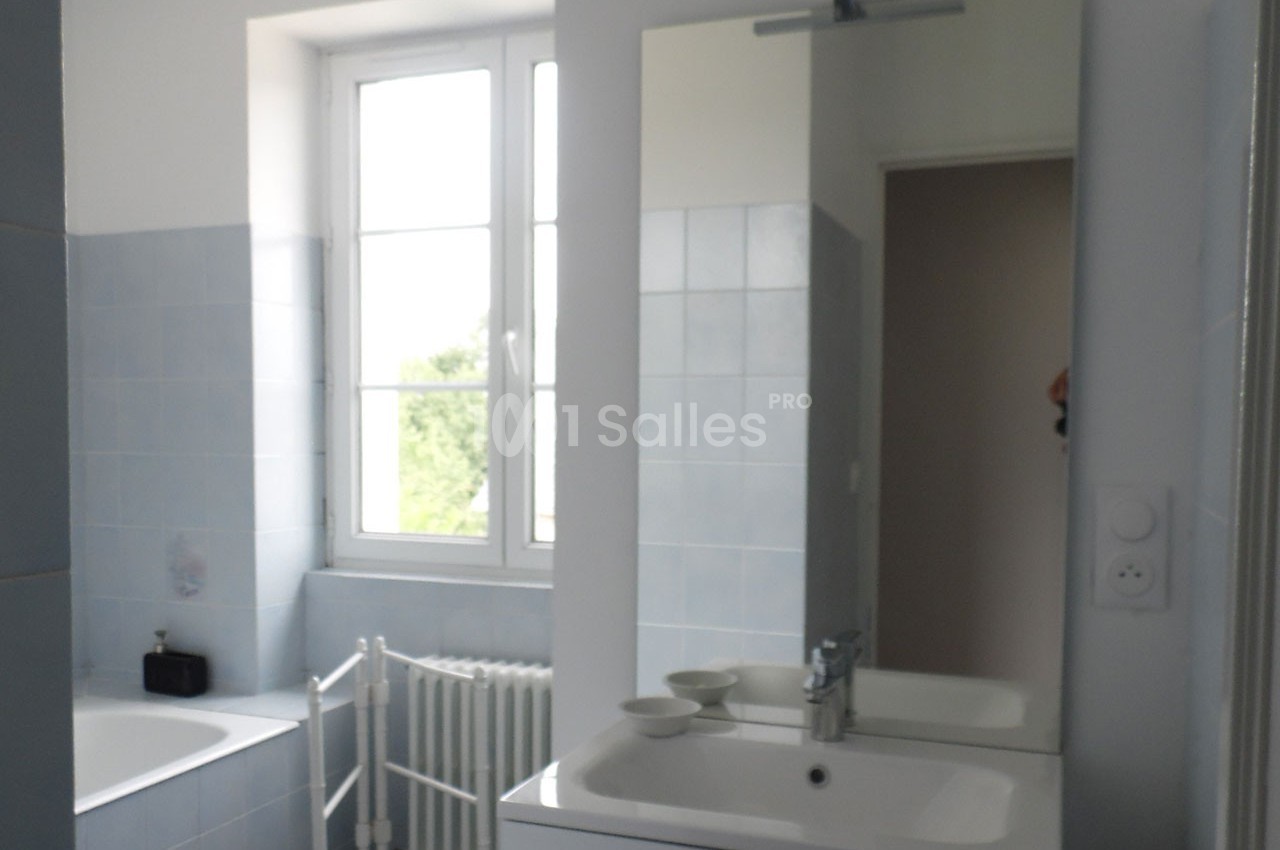Salle de bain lumineuse avec carrelage bleu clair, lavabo, miroir, baignoire et fenêtre donnant sur l'extérieur.