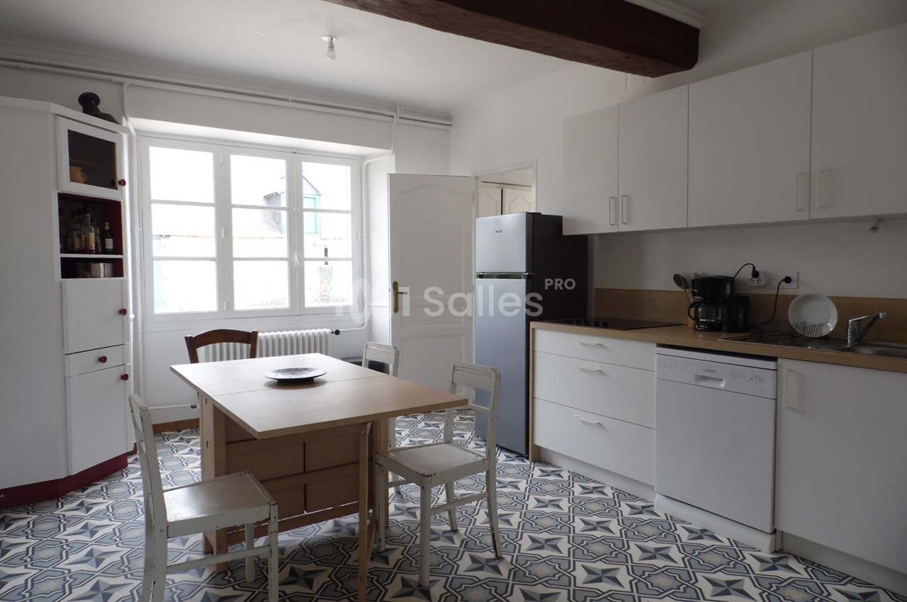 Cuisine lumineuse avec meubles blancs, table en bois, réfrigérateur, lave-vaisselle et sol en carreaux à motifs.