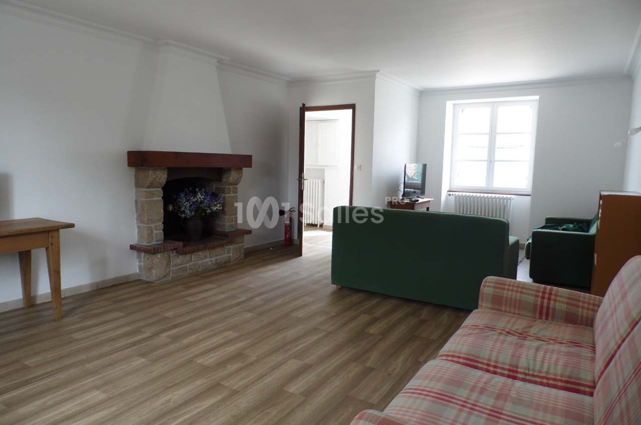 Salon lumineux avec parquet, cheminée en pierre, canapé vert, fauteuil à carreaux et fenêtre laissant entrer la lumière.
