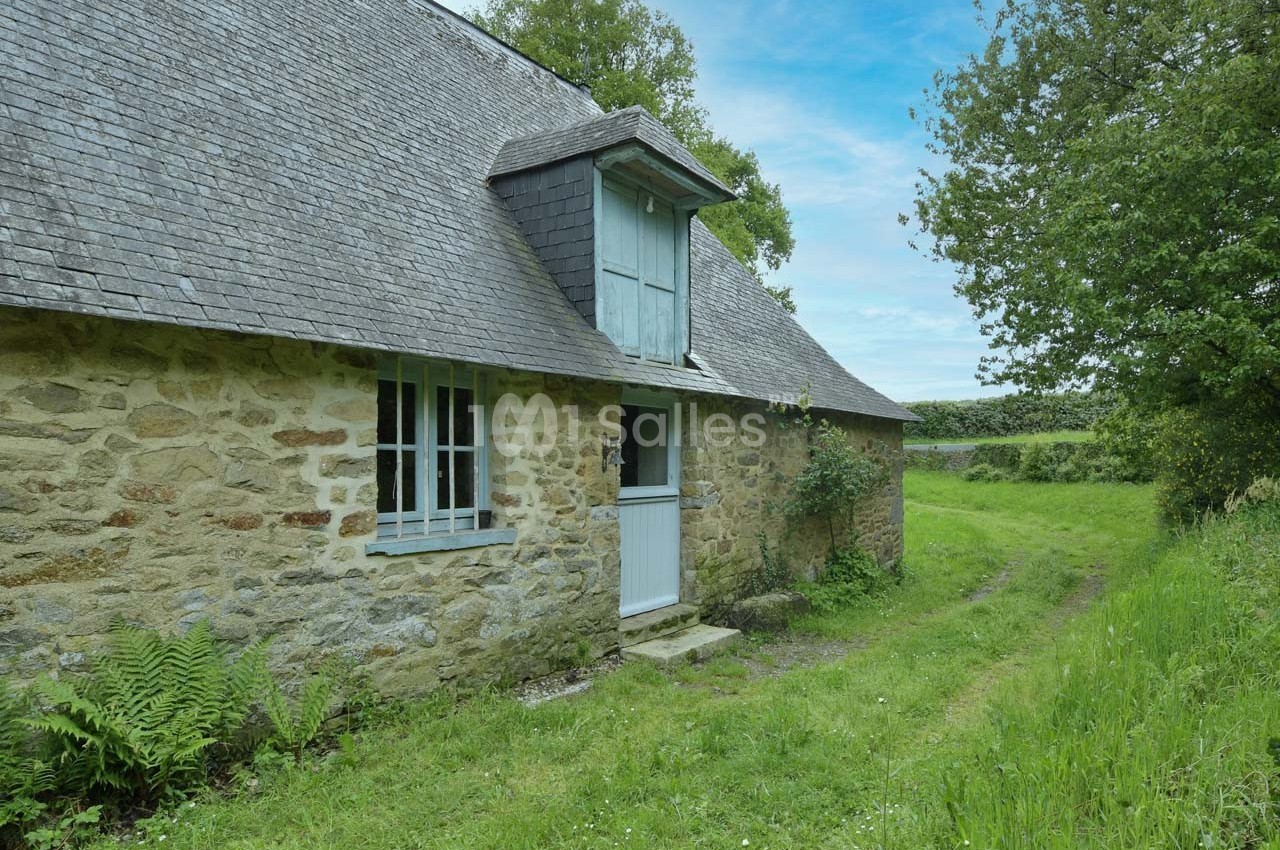 Maison en pierre avec toit en ardoise, entourée de verdure, située le long d'un chemin herbeux.