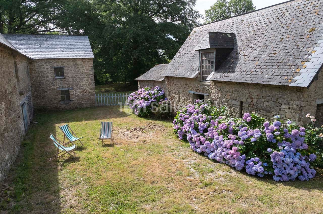 Cour enherbée entourée de bâtiments en pierre, avec des hortensias en fleurs et deux chaises longues colorées.