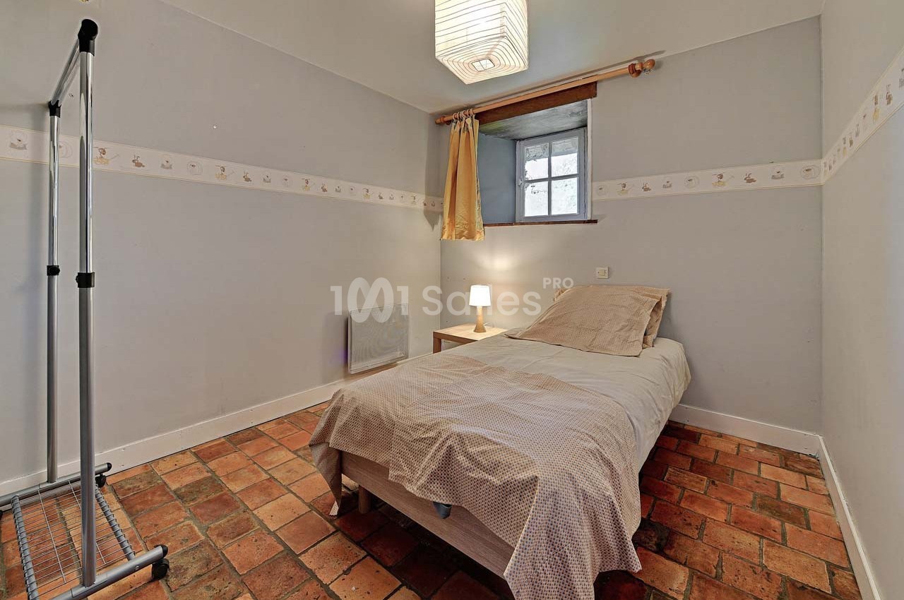 Chambre simple avec lit une place, sol en carreaux de terre cuite, mur gris clair et petite fenêtre avec rideaux.