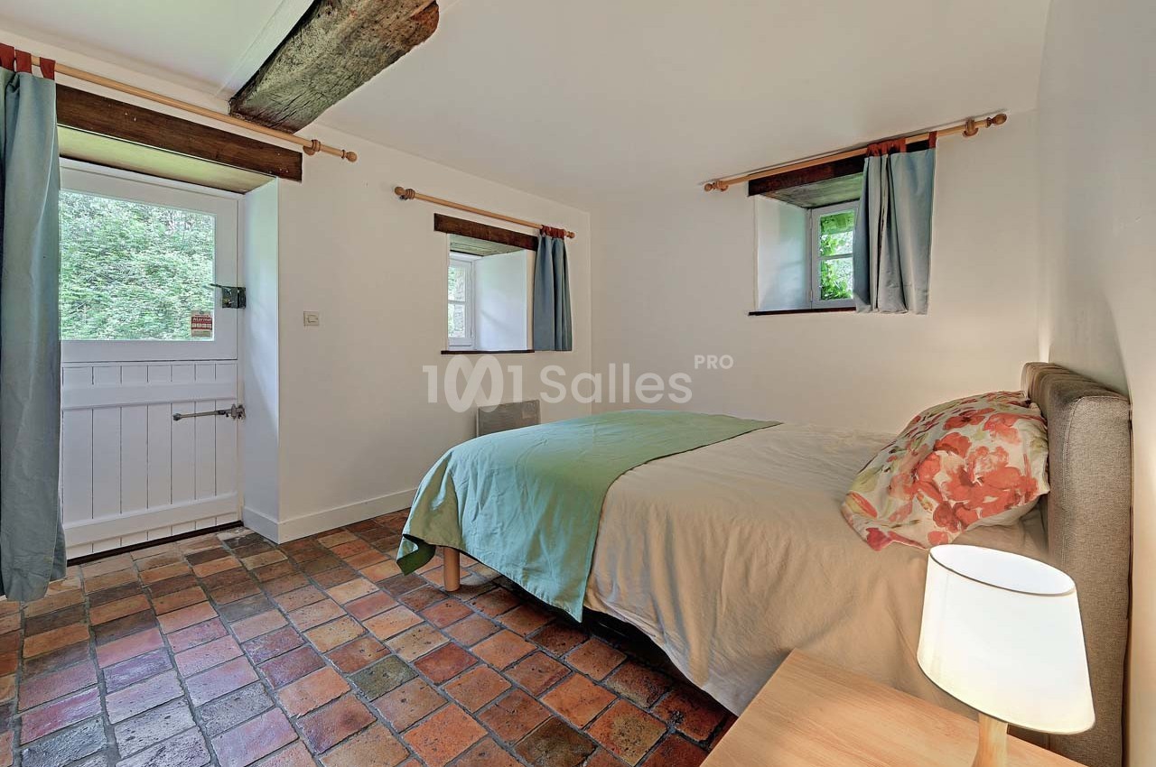 Chambre lumineuse avec lit double, sol en carreaux de terre cuite, poutre apparente et fenêtres avec rideaux bleus.
