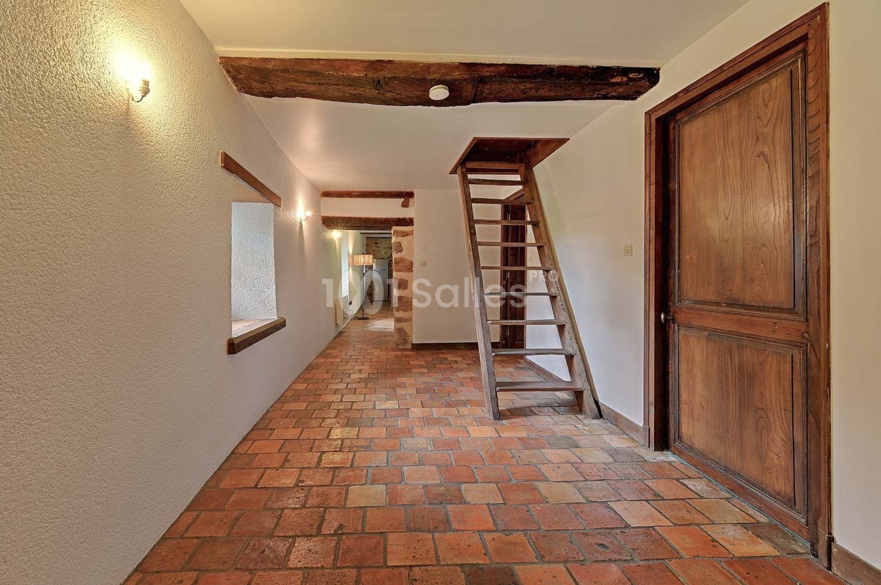 Escalier en bois dans un couloir avec sol en carreaux de terre cuite, murs clairs et poutres apparentes.