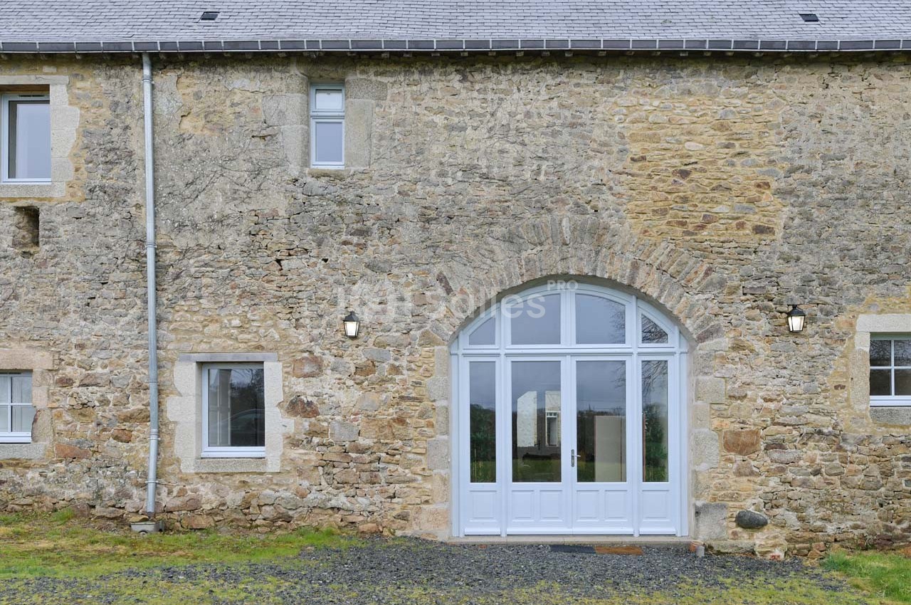 Façade en pierre d'une maison avec une grande porte vitrée cintrée et des fenêtres, entourée de pelouse et de gravier.