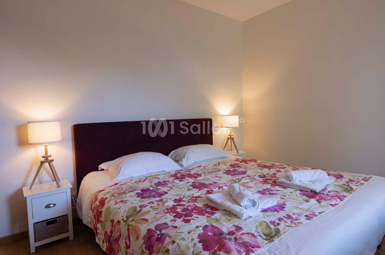 Lit double avec tête de lit violette, drap à motifs floraux, deux tables de chevet blanches et lampes allumées.