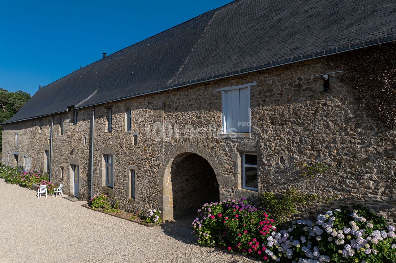 Façade en pierre d'une bâtisse ancienne avec des hortensias colorés et une cour gravillonnée sous un ciel bleu.
