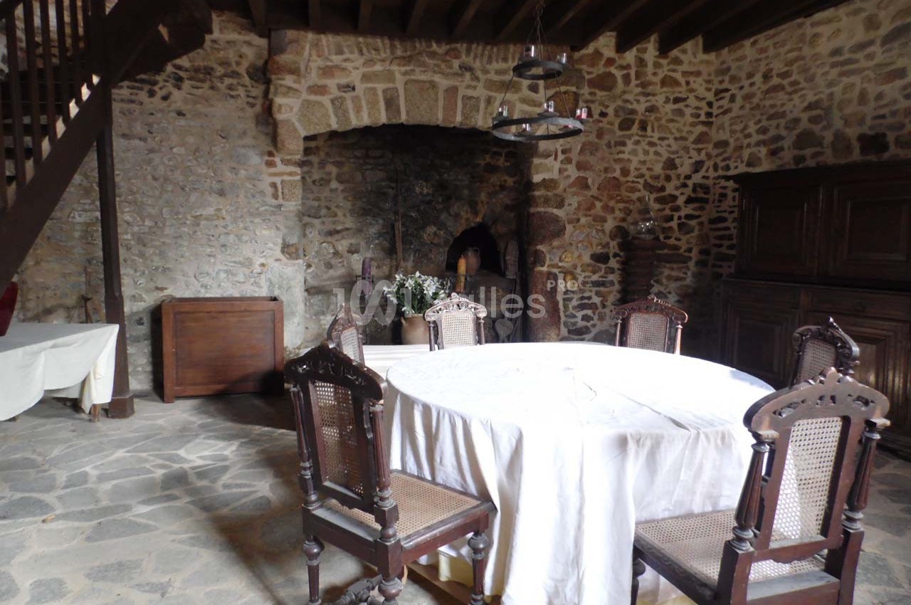 Salle rustique avec murs en pierre, table ronde couverte d'une nappe blanche, chaises en bois et cheminée ancienne.