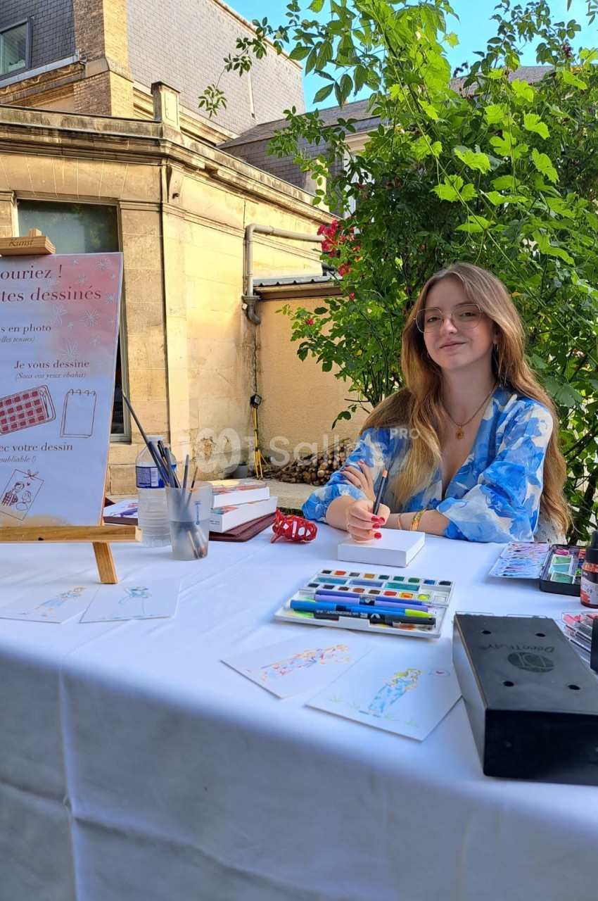 Une artiste assise à une table en extérieur, entourée de matériel de dessin et de peintures.