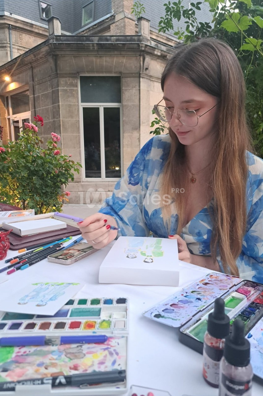 Une femme peint à l'aquarelle sur une table en extérieur, entourée de fournitures artistiques et de végétation.