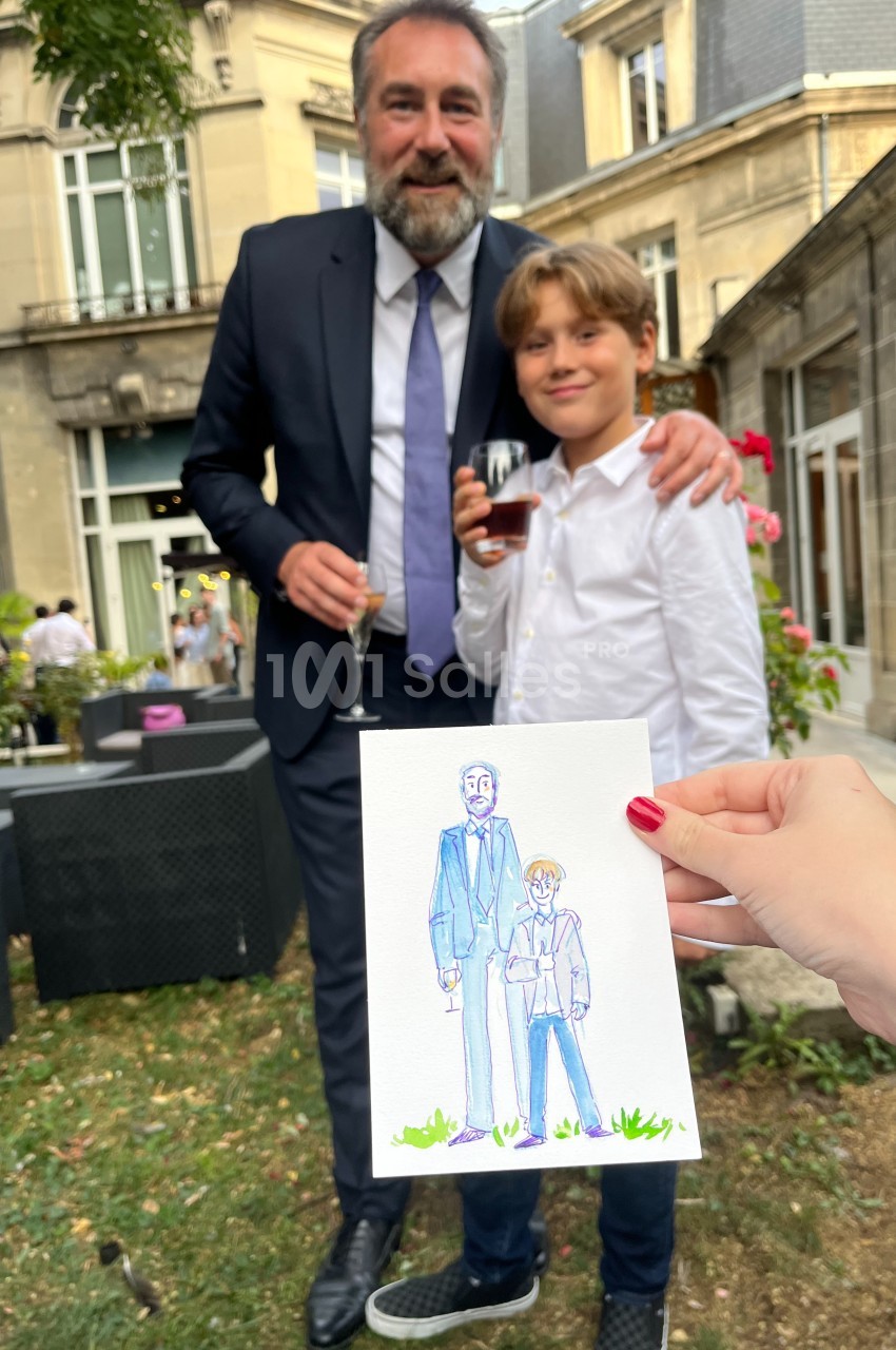 Un homme en costume et un enfant souriant posent dans un jardin, avec un dessin les représentant tenu au premier plan.