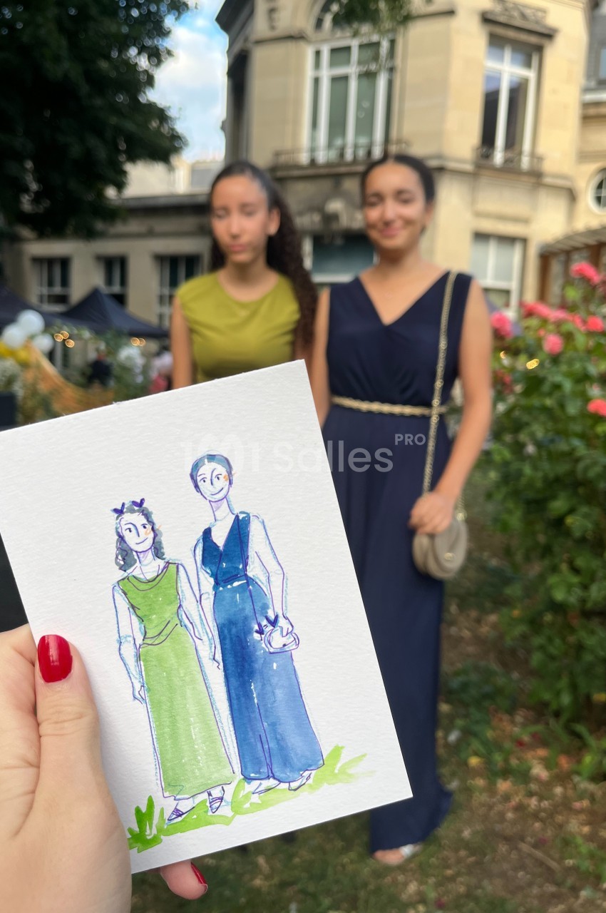 Une main tient un dessin représentant deux femmes posant dans un jardin, avec les modèles en arrière-plan flou.