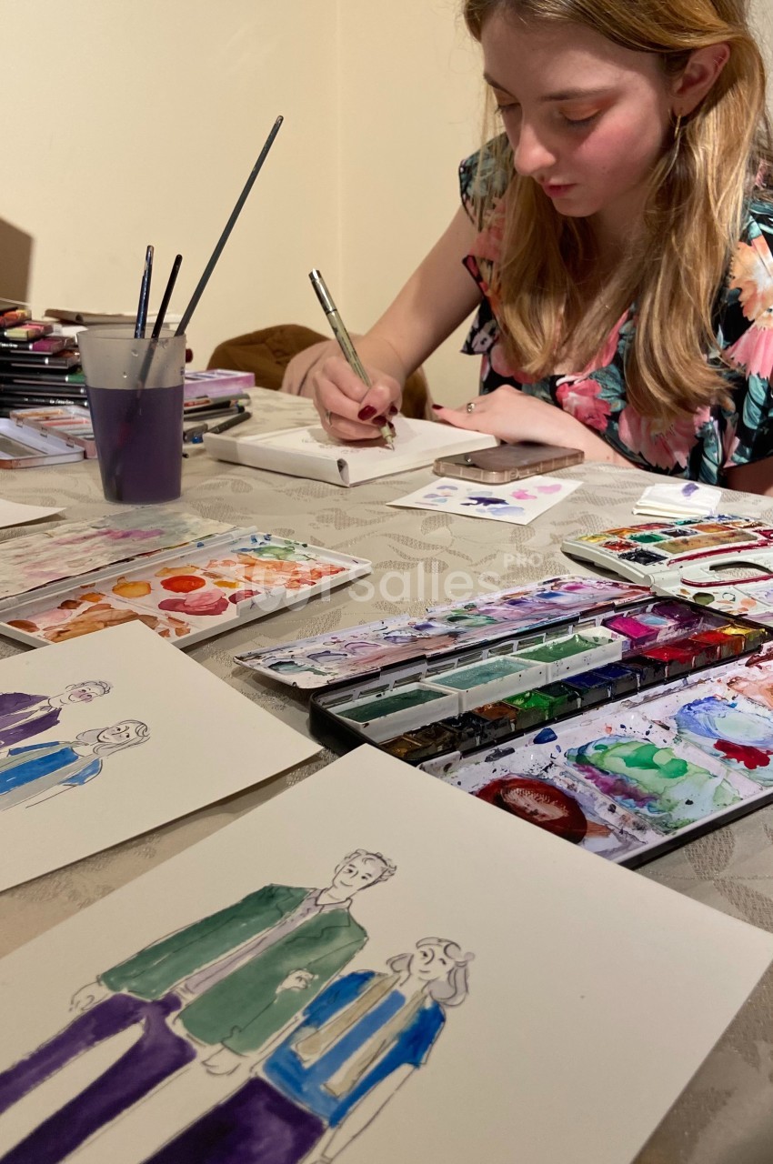 Une jeune femme peint des illustrations colorées à l'aquarelle sur une table remplie de matériel artistique.