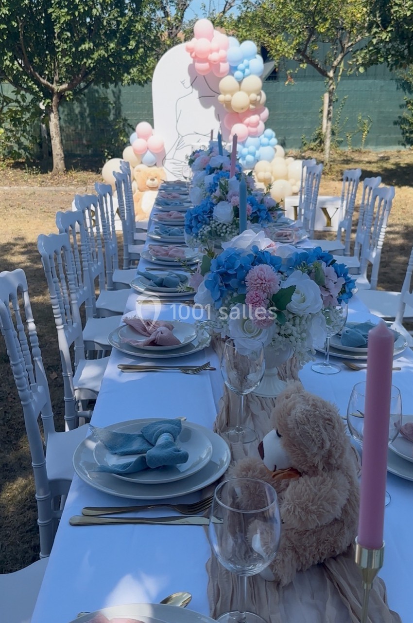 Table décorée pour une fête en extérieur avec fleurs, peluches, bougies roses et ballons en arrière-plan.