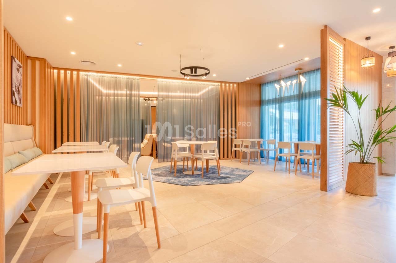 Salle lumineuse avec tables et chaises modernes, décorée de bois clair, plantes et rideaux bleus translucides.