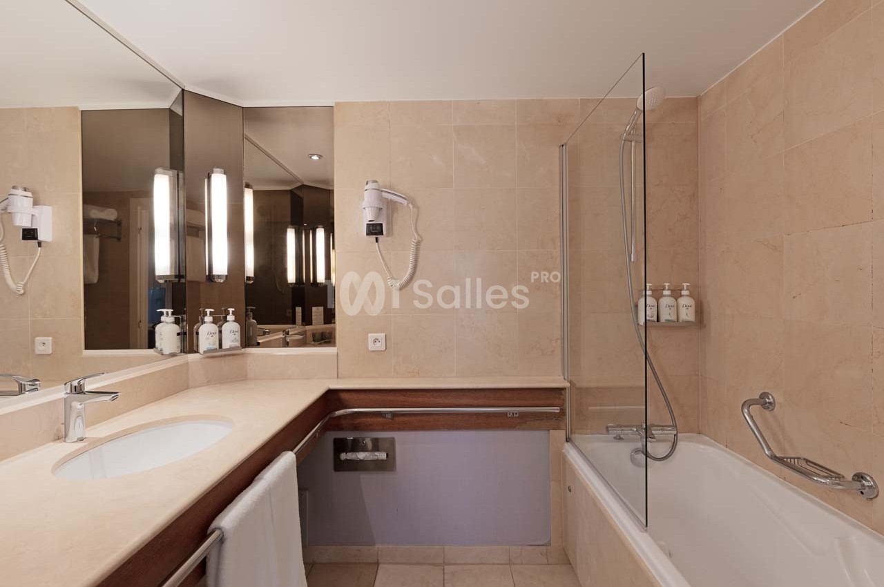 Salle de bain avec baignoire, lavabo, miroir éclairé, sèche-cheveux mural et distributeurs de savon fixés au mur.
