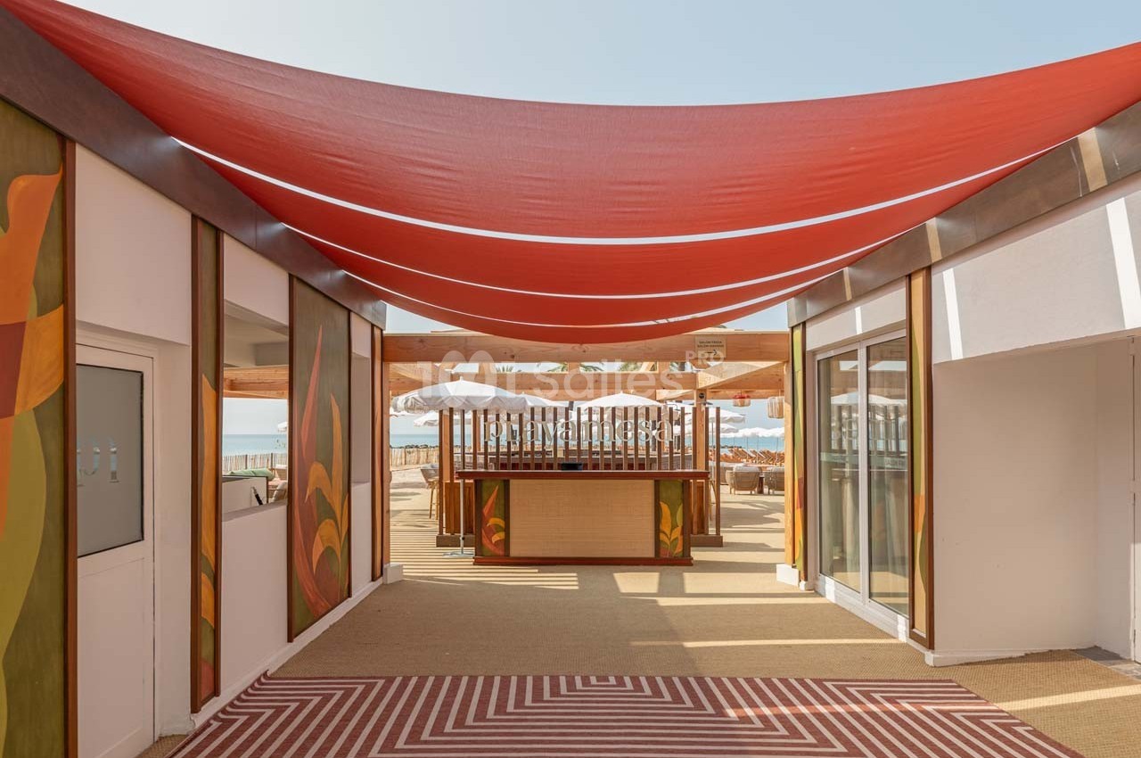 Vue d'une terrasse extérieure avec une toile rouge tendue, des motifs colorés et un bar en arrière-plan.
