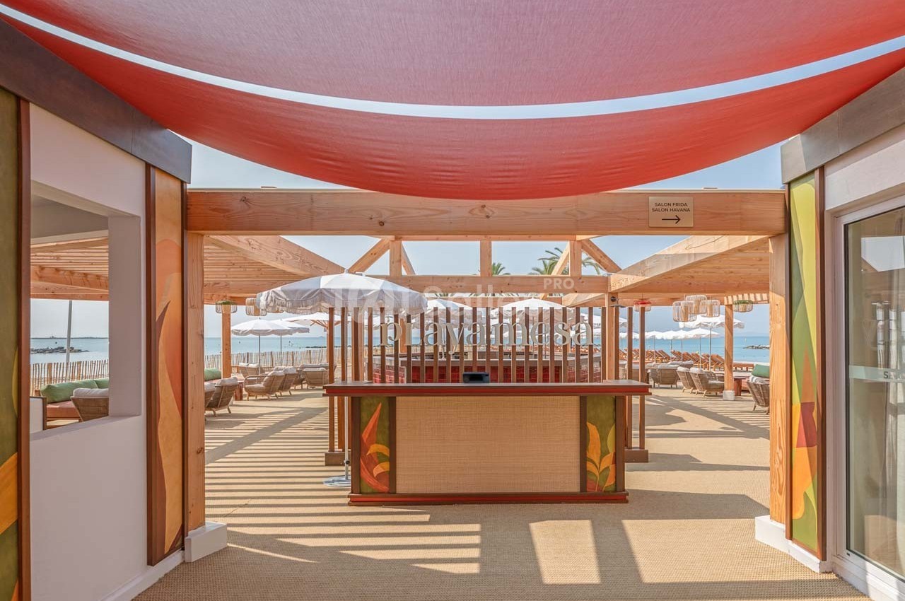 Vue d'une terrasse en bois avec un comptoir central, des parasols blancs et la mer en arrière-plan.