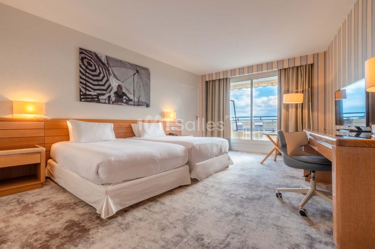 Chambre d'hôtel lumineuse avec deux lits simples, bureau, fauteuil, et balcon offrant une vue sur le ciel.
