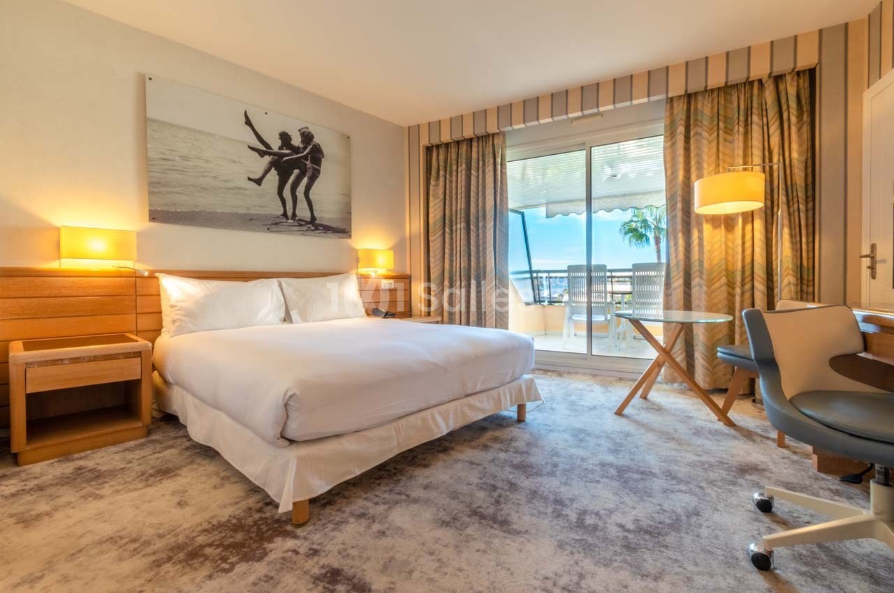 Chambre d'hôtel lumineuse avec lit double, bureau, fauteuil, et accès à un balcon avec vue sur l'extérieur.