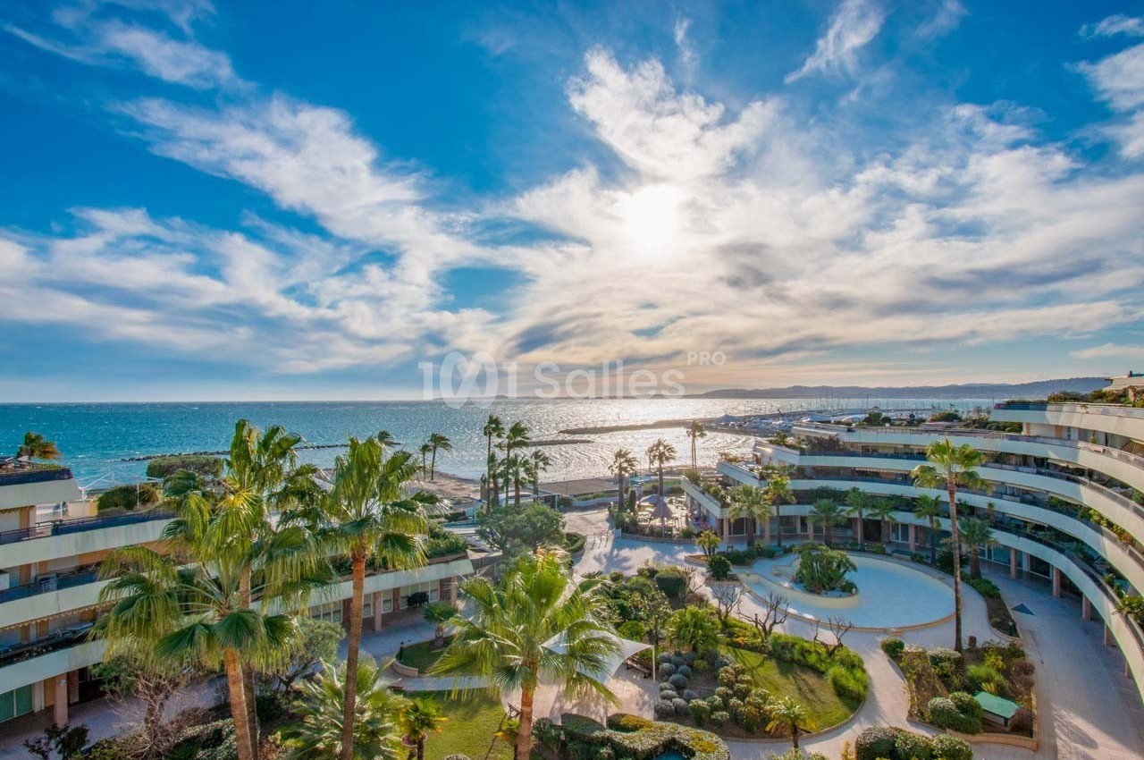 Vue sur une résidence moderne avec jardins, palmiers et en arrière-plan la mer sous un ciel partiellement nuageux.