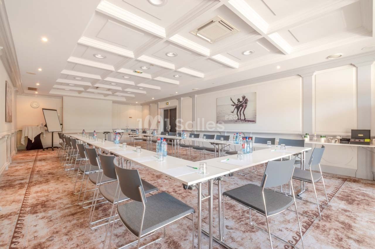 Salle de réunion lumineuse avec tables disposées en U, chaises modernes et équipement de présentation.