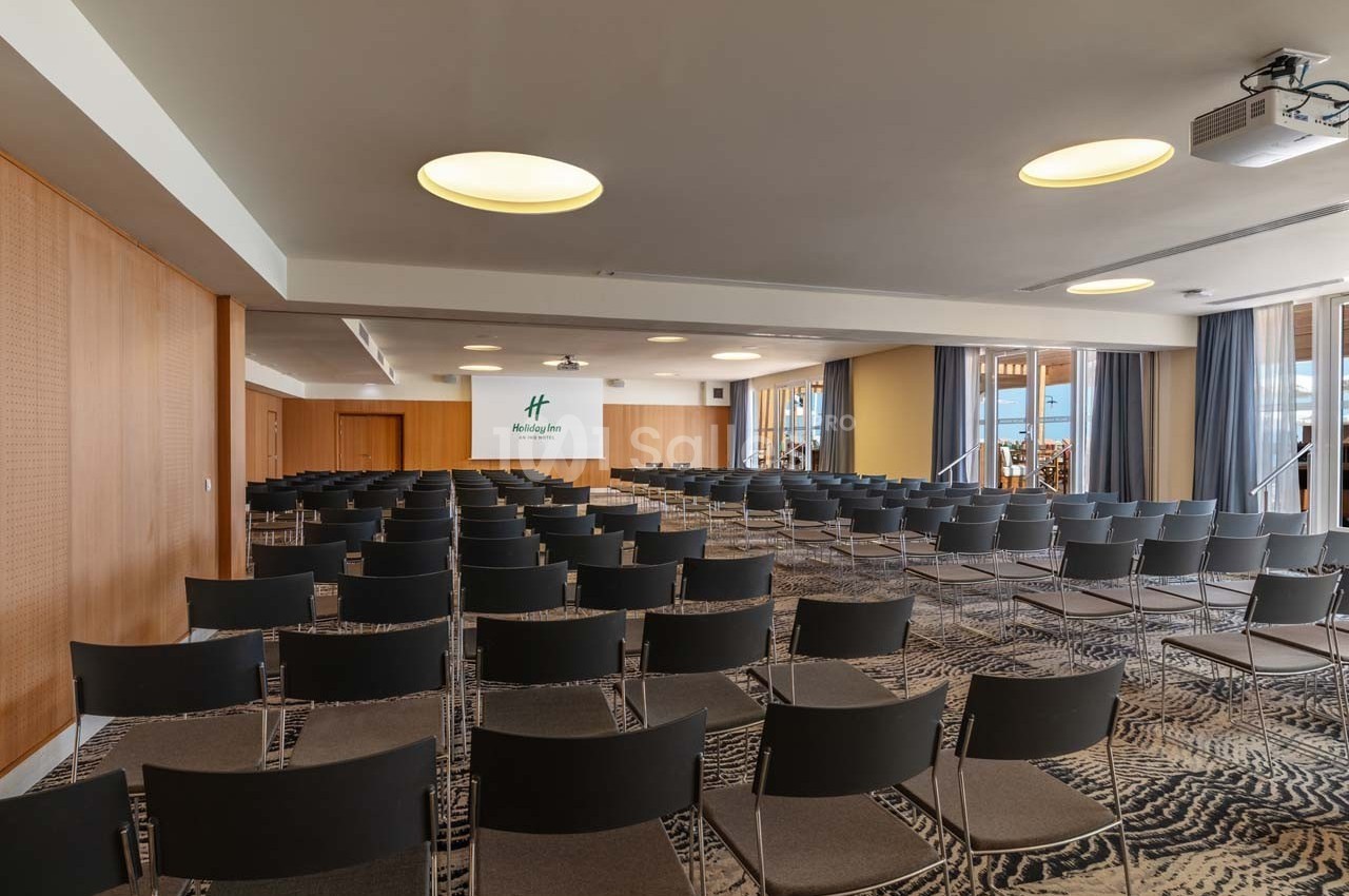 Salle de conférence avec rangées de chaises noires, éclairage moderne et écran de projection au fond.