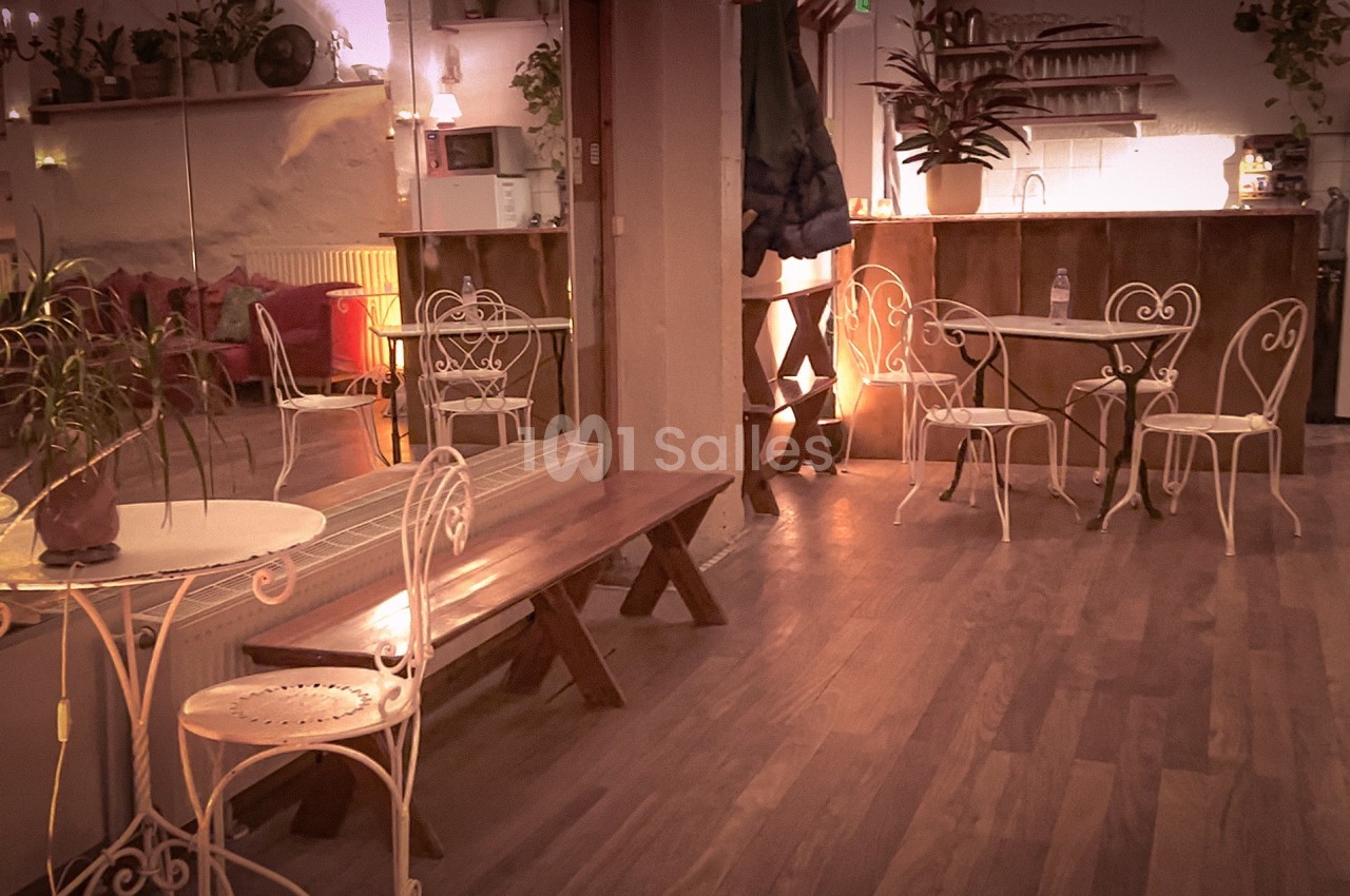 Salle chaleureuse avec tables et chaises en métal blanc, bancs en bois, plantes et éclairage tamisé.