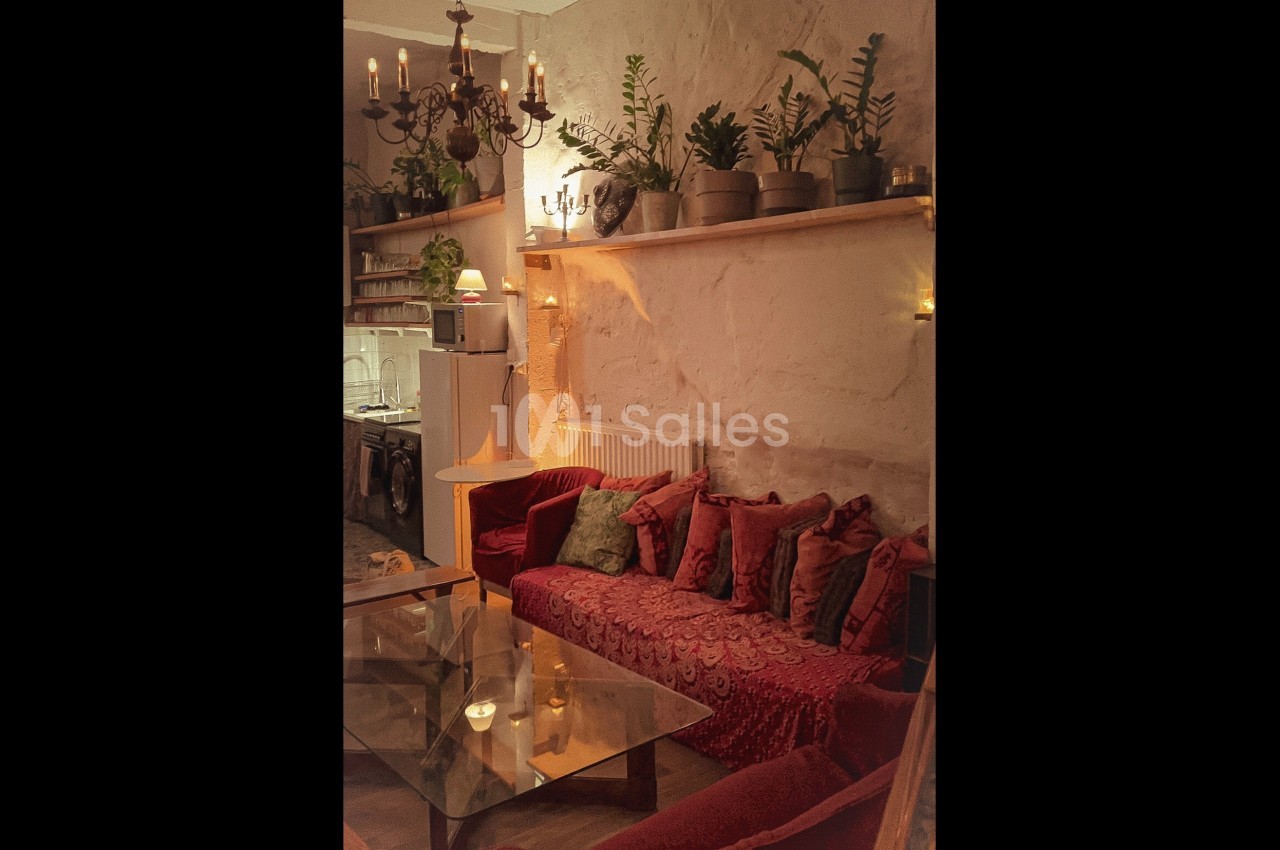 Salon chaleureux avec canapé rouge, table en verre, plantes en pot et éclairage tamisé sous un lustre ancien.