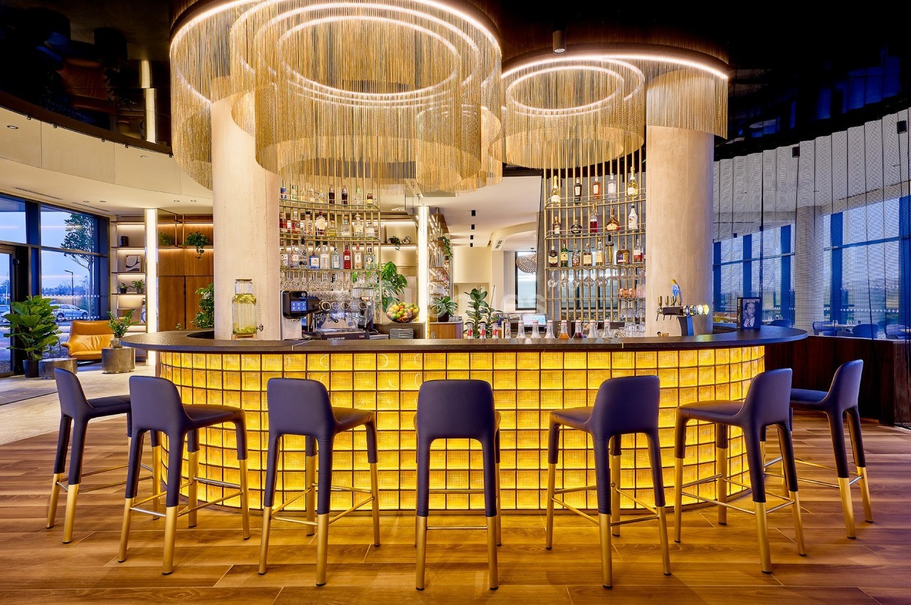 Bar moderne avec comptoir lumineux en verre, tabourets bleus et dorés, et lustres circulaires suspendus.