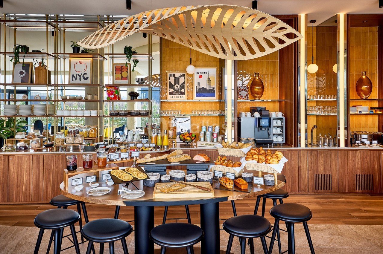 Buffet de petit-déjeuner avec viennoiseries, pains, confitures et boissons dans un espace lumineux et moderne.