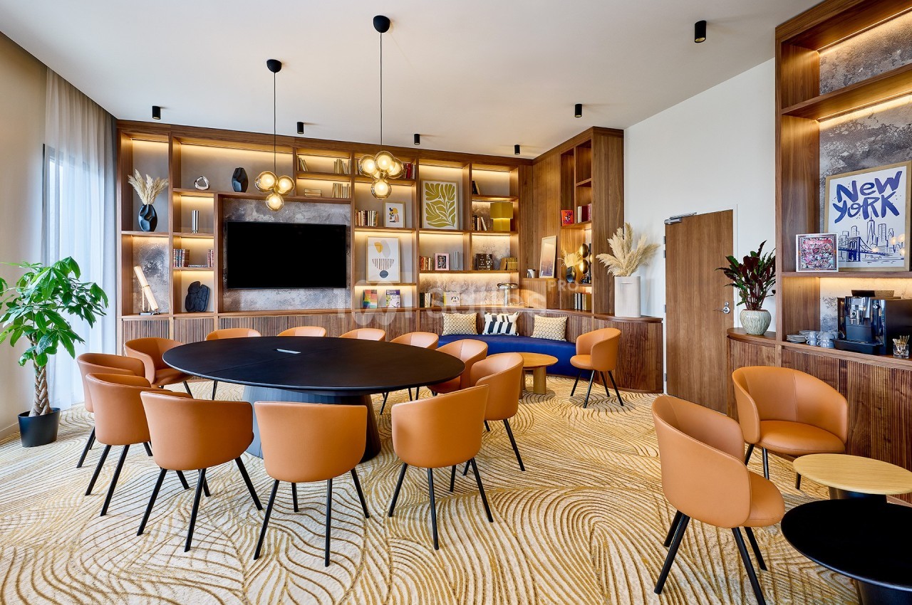 Salle de réunion moderne avec table ronde, chaises en cuir orange, étagères décorées et éclairage suspendu.