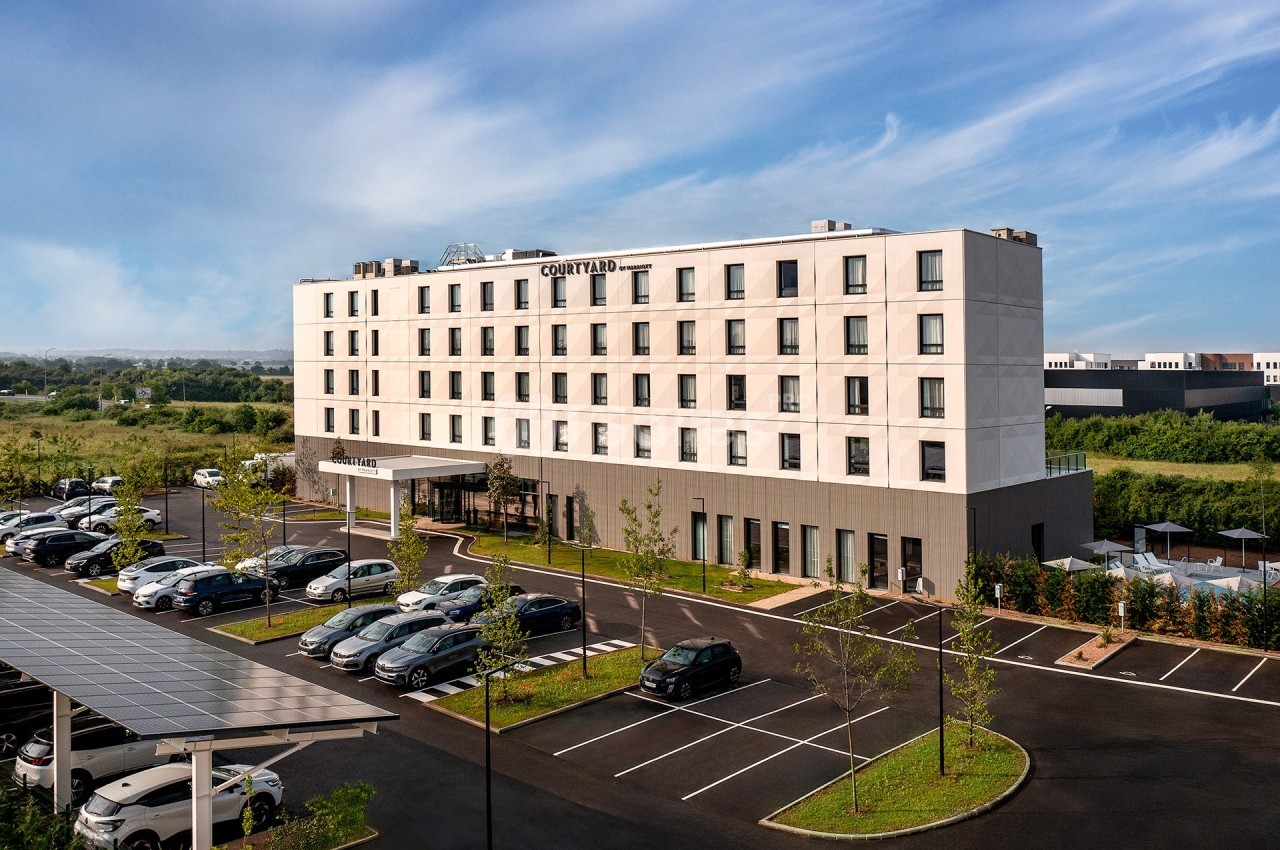 Façade d'un hôtel moderne avec parking extérieur, entouré de végétation et sous un ciel dégagé.