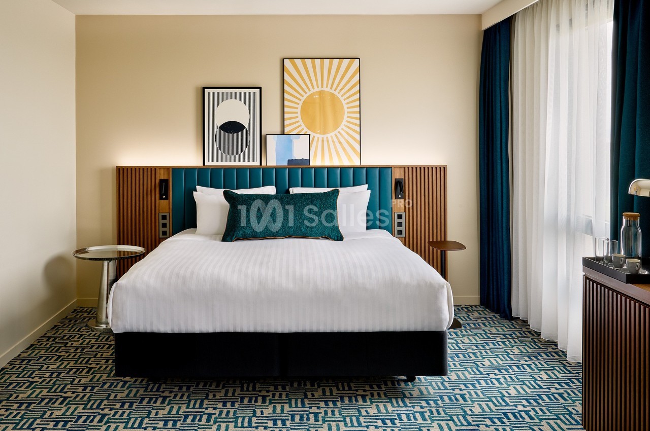 Lit double avec tête de lit bleue, coussin décoratif, tables de chevet et décorations murales dans une chambre lumineuse.