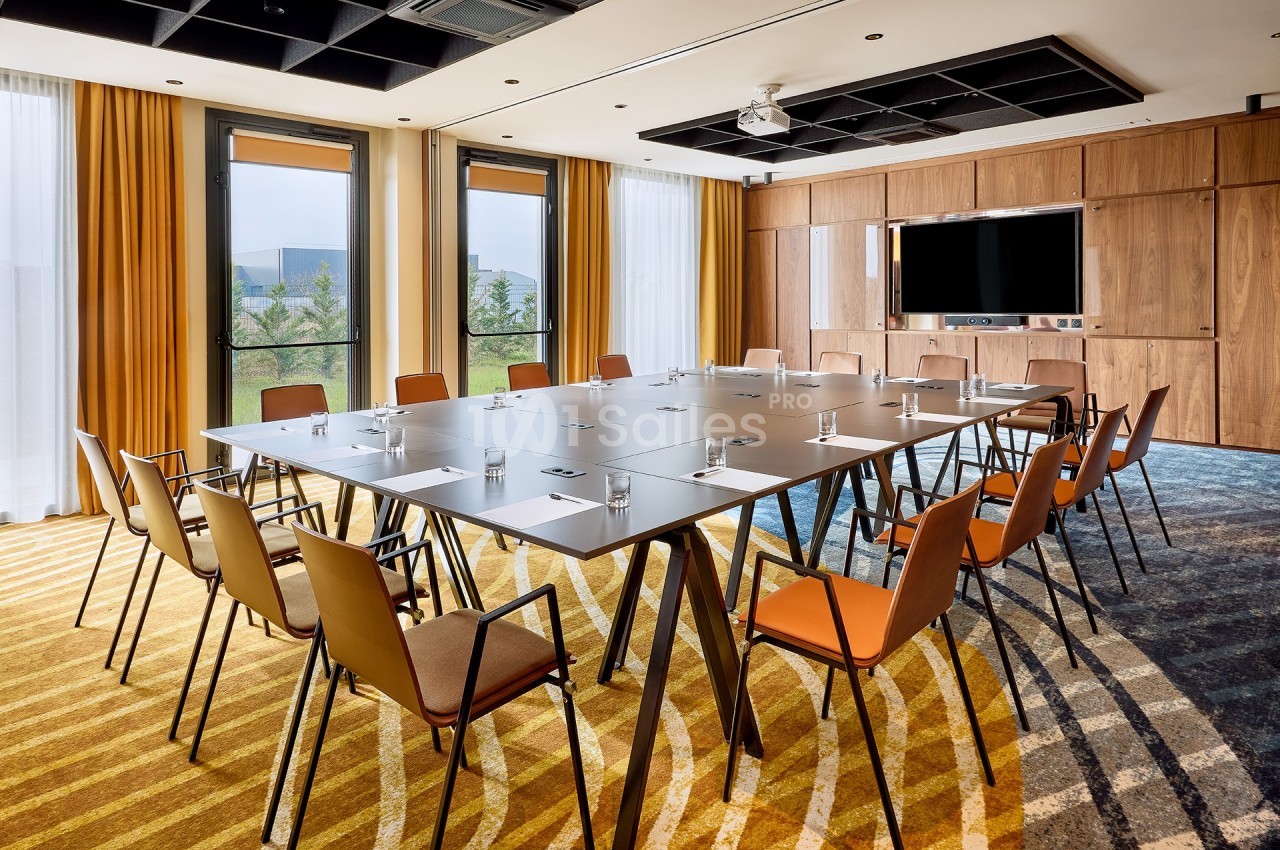 Salle de réunion lumineuse avec une grande table rectangulaire, des chaises orange et un écran mural.