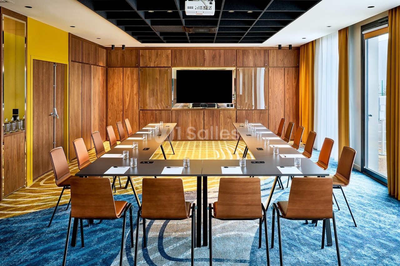 Salle de réunion moderne avec table en U, chaises en bois, écran mural et grandes fenêtres laissant entrer la lumière…