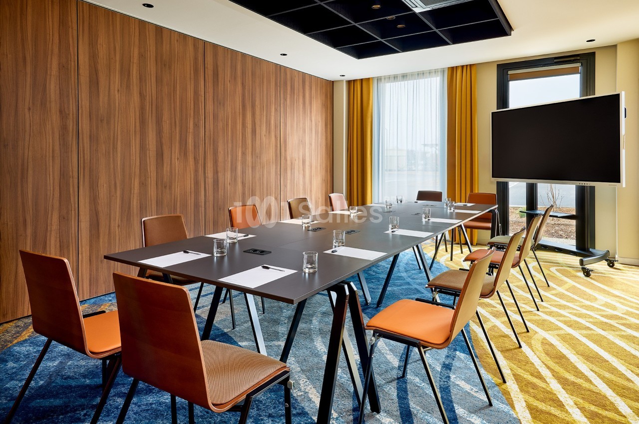 Salle de réunion moderne avec table rectangulaire, chaises orange, écran sur pied et lumière naturelle.