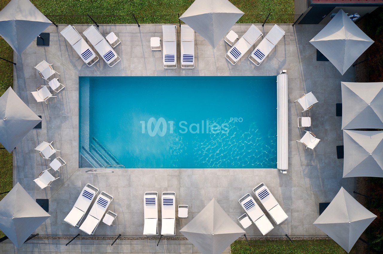 Vue aérienne d'une piscine rectangulaire entourée de transats blancs, de parasols et d'une terrasse en dalles grises.